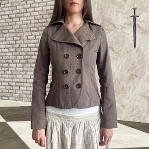 Puede incluir: Una chaqueta cruzada de color marrón con un sutil patrón de cuadros, cuello ancho y seis botones oscuros dispuestos en dos filas verticales. La chaqueta tiene mangas largas y un corte entallado, sobre una blusa blanca y una falda clara con volantes.