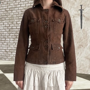 Könnte beinhalten: Eine dunkelbraune, taillierte Jacke mit strukturiertem Kragen, langen Ärmeln und einer durchgehenden Knopfleiste. Das Kleidungsstück verfügt über zwei Brusttaschen und zwei untere Taschen, die jeweils mit einem Metallknopf versehen sind.