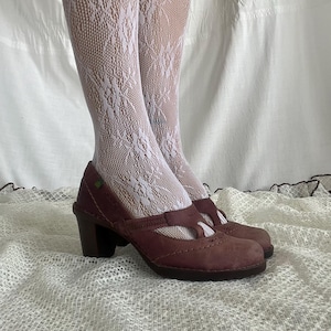Op de afbeelding: Een paar donkerkastanjebruine leren mary jane pumps met een ronde neus, een dikke blokhak en zichtbare decoratieve stiksels langs de wreef en zijkanten. De schoenen worden gedragen met een witte kanten panty met een gedetailleerd bloemenpatroon.