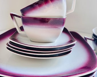May include: A set of square ceramic dishes and cups featuring a white center that transitions into a deep purple gradient along the edges. The cups have angular handles and are stacked on top of the square saucers and larger square plates.
