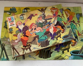 May include: A vintage 60-piece puzzle featuring a scene of seven dwarfs sitting at a table with a pie, surrounded by a forest setting. The puzzle box is labeled "MB Puzzle 60 Pieces".