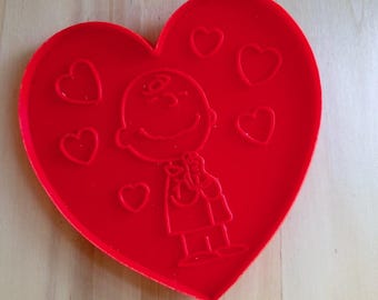 May include: A bright red heart-shaped cookie cutter featuring an embossed design of a cartoon character with a round head and simple facial features, surrounded by five small heart outlines on the surface of the red plastic material.