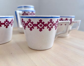 May include: A white ceramic tea set with a maroon geometric pattern and blue trim. The set includes cups and a teapot. The cups have square handles. The tea set is on a light-colored wooden surface.