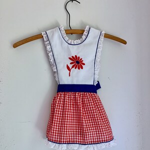 May include: White sleeveless dress with a red flower embroidered on the bodice, a blue sash, and a red and white gingham skirt.