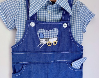 May include: A blue denim overall set with a matching blue and white gingham shirt. The overalls feature a wagon applique and white stitching. The shirt has short sleeves and a button-down front. The outfit is displayed on a hanger.