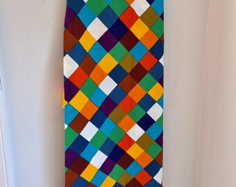 May include: A rectangular fabric piece featuring a repeating diamond pattern in vibrant colors including orange, purple, white, blue, green, yellow, and dark red. The textile is draped over a black hanger, displaying a consistent geometric grid design.