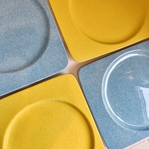 May include: Four square ceramic plates, two are yellow with a raised circular rim and two are blue with a raised circular rim and a white swirl design.