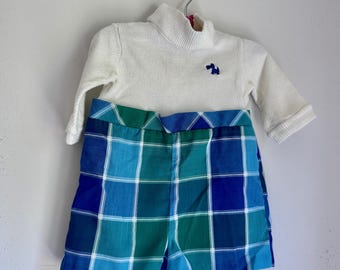 May include: A vintage baby outfit featuring a white ribbed turtleneck sweater with a small blue embroidered design and plaid shorts in shades of blue and green. The outfit is displayed on a hanger.