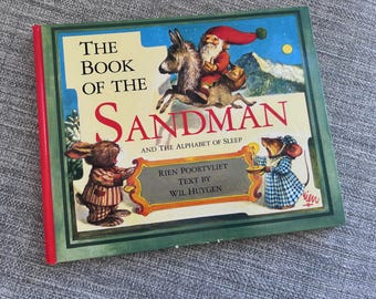 May include: A hardcover children's book titled "The Book of the Sandman and the Alphabet of Sleep." The cover features illustrations of a gnome riding a donkey, a rabbit, and a mouse. The book has a green border and a red spine.