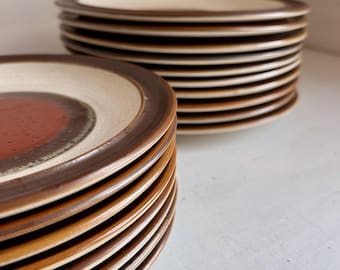 May include: A stack of round ceramic plates featuring a dark reddish-brown center, a wide cream-colored band, and a dark brown rim. The plates are arranged in two separate piles, showcasing the layered design and the smooth, glazed finish.
