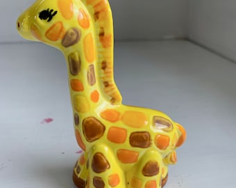 May include: A yellow ceramic giraffe figurine with brown spots. The giraffe has a long neck and a small head with a black eye.