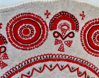 May include: Close-up of a white linen tablecloth with red and black embroidery. The embroidery features a circular design with concentric circles, a floral motif, and a zig-zag pattern.
