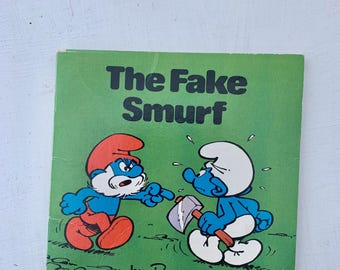 May include: A green book cover with the title "The Fake Smurf" in black text. The cover features two Smurfs, one with a red hat and a beard, and the other with a white hat. The book is by Peyo.