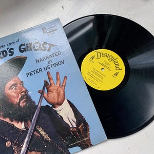 May include: A vintage vinyl record with a black label and a yellow label that says "Disneyland" and "The Story of Blackbeard's Ghost". The record is on top of a blue and white album cover with a picture of a man with a beard and a sword.