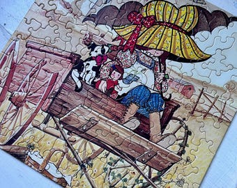 May include: A completed jigsaw puzzle featuring a pastoral scene. The image depicts a person in a yellow hat and blue pants, seated in a wooden cart, holding an umbrella. Two dogs and a child are also in the cart. The puzzle has a beige background.