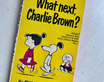 May include: A vintage paperback book titled "What next, Charlie Brown?" with a bright yellow cover. The cover features illustrations of Charlie Brown, Snoopy, and other Peanuts characters. The book is by Charles M. Schulz.