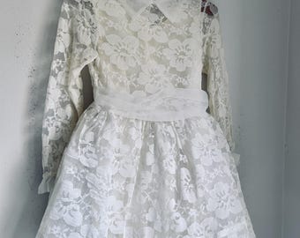 May include: A white lace dress with long sleeves and a Peter Pan collar hangs on a wooden hanger. The dress features a fitted bodice, a defined waist, and a flared skirt. The delicate floral lace pattern covers the entire dress.