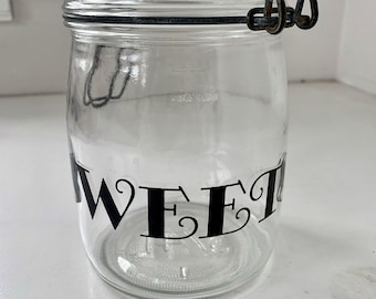 May include: A clear glass jar with a black metal lid and a wire closure. The jar has the word "SWEET" written on the front in black lettering.