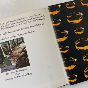 May include: An open book with a black and yellow cover featuring a repeating pattern of gold rings. The book is open to a page with an illustration of two figures walking through a forest. The text on the page reads "Here ends the first part of the History of the War of the Ring."