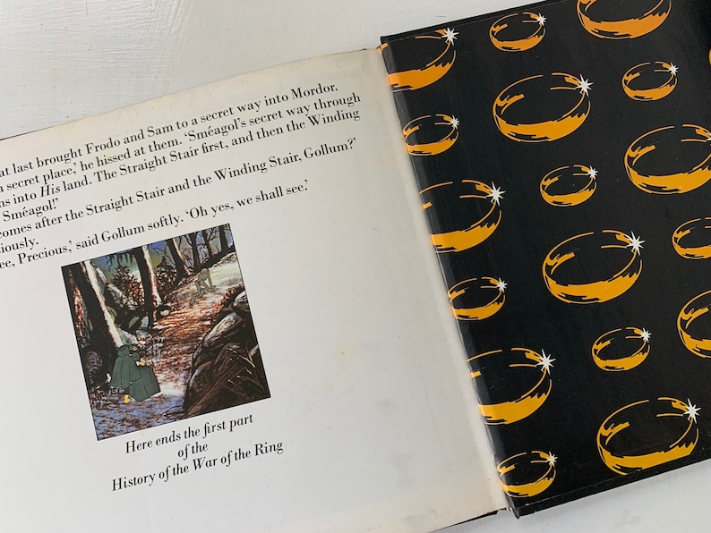 May include: An open book with a black and yellow cover featuring a repeating pattern of gold rings. The book is open to a page with an illustration of two figures walking through a forest. The text on the page reads "Here ends the first part of the History of the War of the Ring."