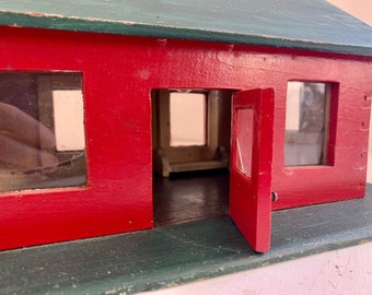 May include: A red wooden toy house with a green roof and a door that is open. The house has three windows, one of which is a door. The house is sitting on a green base.