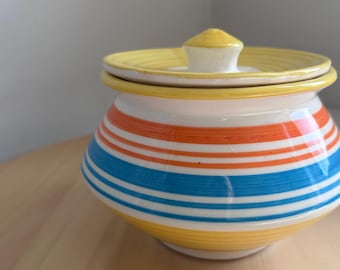 May include: A ceramic jar with a lid, featuring horizontal stripes in orange, blue, and yellow against a white background. The lid and base are yellow. The jar has a small, white knob on top.