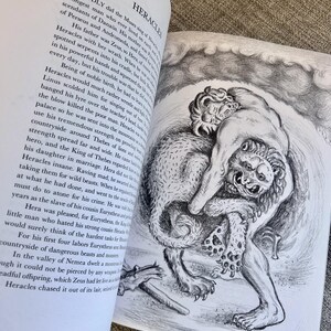 以下が含まれることがあります： ヘラクレスと題された白黒の印刷テキストと、筋肉質の人物が大きなライオンのような生き物と格闘している詳細な鉛筆画が描かれた開いた本のページ。背景には渦巻く雲が描かれ、格闘の様子が表現されています。