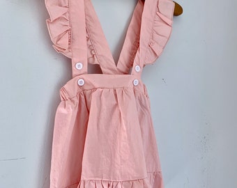 May include: A pink sleeveless dress with a ruffled neckline and straps. The dress has a gathered skirt with a ruffle at the hem and two white buttons on the straps.