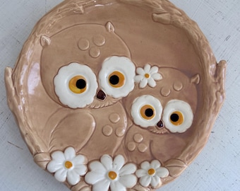 May include: A ceramic owl-themed decorative plate. The plate features two owls with large, white-rimmed eyes and yellow and black pupils. White daisies with yellow centers are placed around the owls. The plate is a light brown color.