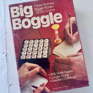May include: A red box game called "Big Boggle" by Parker Brothers. The box shows a close-up of the game board with letter cubes and a person writing down words on a notepad. The text on the box says "50% More Cubes, Longer Words, Greater Challenge".