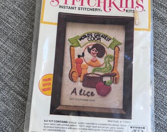 May include: A vintage Stitchkins embroidery kit featuring a framed design that reads "World's Greatest Cook." The kit includes a 5x7 inch design with the name "Alice" and a red apple. The packaging is yellow and white.