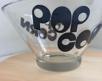 May include: Clear glass popcorn bowl with the word "popcorn" in black lettering. The bowl has a wide, flared shape and a sturdy base. The text is in a bold, rounded font.