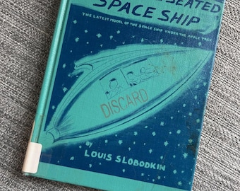 May include: A hardcover book titled The Three-Seated Space Ship by Louis Slobodkin. The cover is teal and dark blue with a graphic of a space ship and stars. The word DISCARD is printed in orange across the center of the space ship illustration.