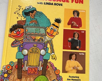 May include: A yellow hardcover book titled Sesame Street Sign Language Fun with Linda Bove. The cover features Cookie Monster, Bert, and Ernie in a purple car, a green turtle, and three photographs of a person demonstrating signs for cookie, car, and turtle.