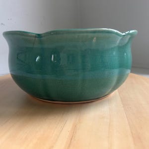 May include: A teal ceramic bowl with a scalloped rim and a visible crackle glaze finish. The bowl features a rounded body with subtle vertical indentations along the sides, creating a fluted appearance. The base is a light, unglazed clay color.