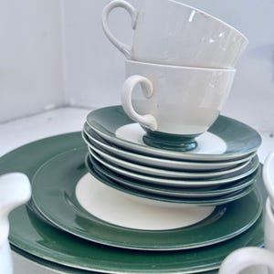 May include: A stack of white teacups with curved handles sits atop a collection of green and white plates. The plates are stacked in a neat pile, with a larger plate at the bottom. The teacups and plates are part of a tea set.