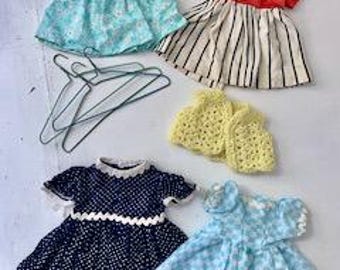 May include: Five doll dresses, including a blue and white floral dress, a red and white striped dress, a yellow crocheted vest, a navy blue and white polka dot dress, and a light blue and white checkered dress.  Four green wire hangers are also included.