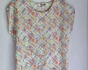 May include: A light yellow, short-sleeved blouse with a geometric pattern in shades of pink, blue, and purple. The blouse has a round neckline and the brand name "Ecruelle" is printed on the label.