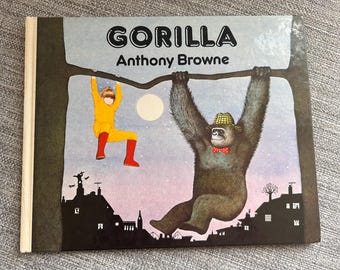May include: A hardcover children's book titled Gorilla by Anthony Browne. The cover illustration depicts a child in a yellow outfit and red boots hanging from a tree branch next to a large gorilla wearing a plaid hat and a red polka dot bow tie.