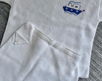 May include: White knitted baby sweater and shorts set with a blue and white embroidered boat design. The sweater has a boat neck and a button closure at the shoulder. The shorts have a pocket on the back.