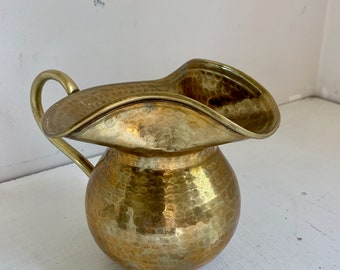 May include: A hammered brass pitcher with a wide, flared lip and a single handle. The pitcher has a round body and a textured surface.