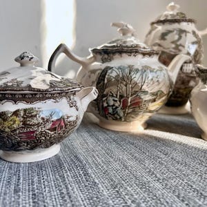 May include: A ceramic tea set featuring a sugar bowl with a lid, a teapot with a lid, and a creamer. Each piece is off-white with a detailed brown and multi-colored landscape transfer print depicting trees, buildings, and a red barn scene.