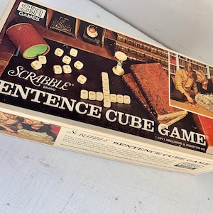 May include: A vintage Scrabble Sentence Cube Game box with a brown and white design. The box features the Scrabble logo and the text "Sentence Cube Game". The box is open and shows the game pieces, which are small cubes with letters on them.
