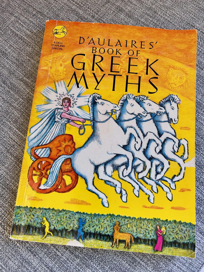 以下が含まれることがあります： 黄色い表紙のペーパーバック「D'Aulaires' Book of Greek Myths」。表紙には、4頭の白い馬に引かれた戦車に乗る人物が描かれており、下部には小さな人物やケンタウロスが風景の中に配置されています。