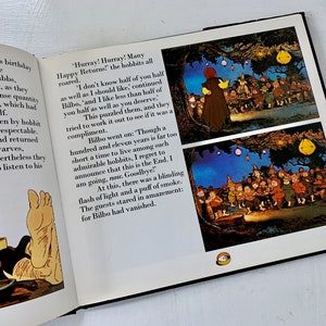 May include: A book open to a page with illustrations of hobbits celebrating a birthday party. The text on the page reads, "Hurray! Hurray! Many Happy Returns!" and "I don't know half of you half as well as I should like, continued Bilbo, 'and I like less than half of you half as well as you deserve."