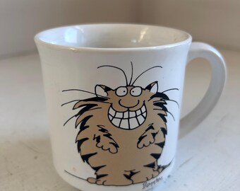 May include: White ceramic mug featuring a cartoon cat with a large, toothy grin. The cat is brown with black stripes and whiskers. The mug has a curved handle and a simple design, suitable for coffee or tea.