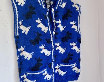 May include: A blue knitted vest with a pattern of white and black dog silhouettes. The vest has a button-up front, two pockets, and white trim along the edges. The vest is hanging on a wooden hanger.