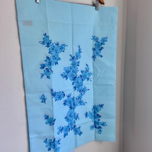 May include: A light blue floral patterned fabric hanging from a wooden hanger. The fabric features clusters of blue flowers in varying shades. The background is a plain white wall.