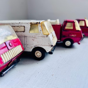 May include: A collection of vintage toy vehicles, including a white camper van and several red and yellow trucks. The toys have a distressed appearance, with visible wear and tear, suggesting they are well-loved.