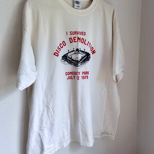 May include: A white short-sleeve t-shirt featuring a black graphic of a baseball stadium and red text that reads I SURVIVED DISCO DEMOLITION COMISKEY PARK JULY 12 1979. The shirt is displayed on a wooden hanger against a plain white wall background.